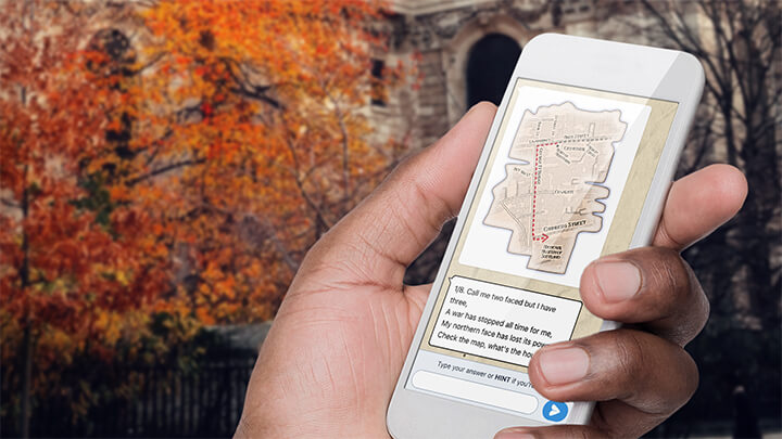 A hand holding a phone playing Treasure Hunt Edinburgh with a tree with orange leaves behind.