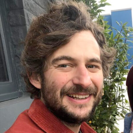 A man with wavy brown hair and a beard, wearing a red jacket, smiling at the camera.